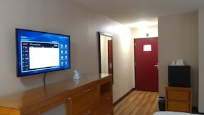 Habitación de hotel con tocador de madera, televisor de pantalla plana mostrando una guía de canales, puerta roja y una mesita con una caja negra.