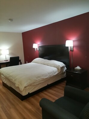 A hotel room with a large bed, two bedside lamps, a desk with a chair, and a dark-colored armchair.