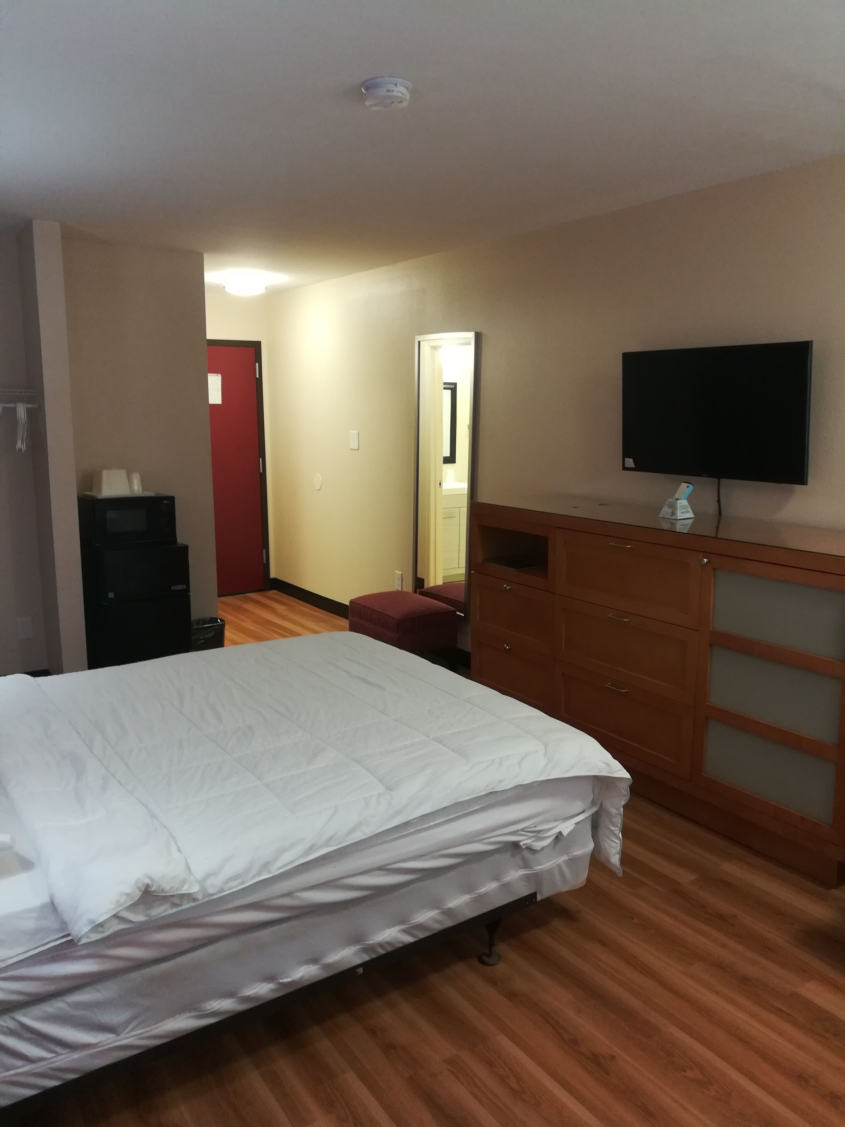 A hotel room with a bed, a wooden dresser, a television mounted on the wall, and a red door.