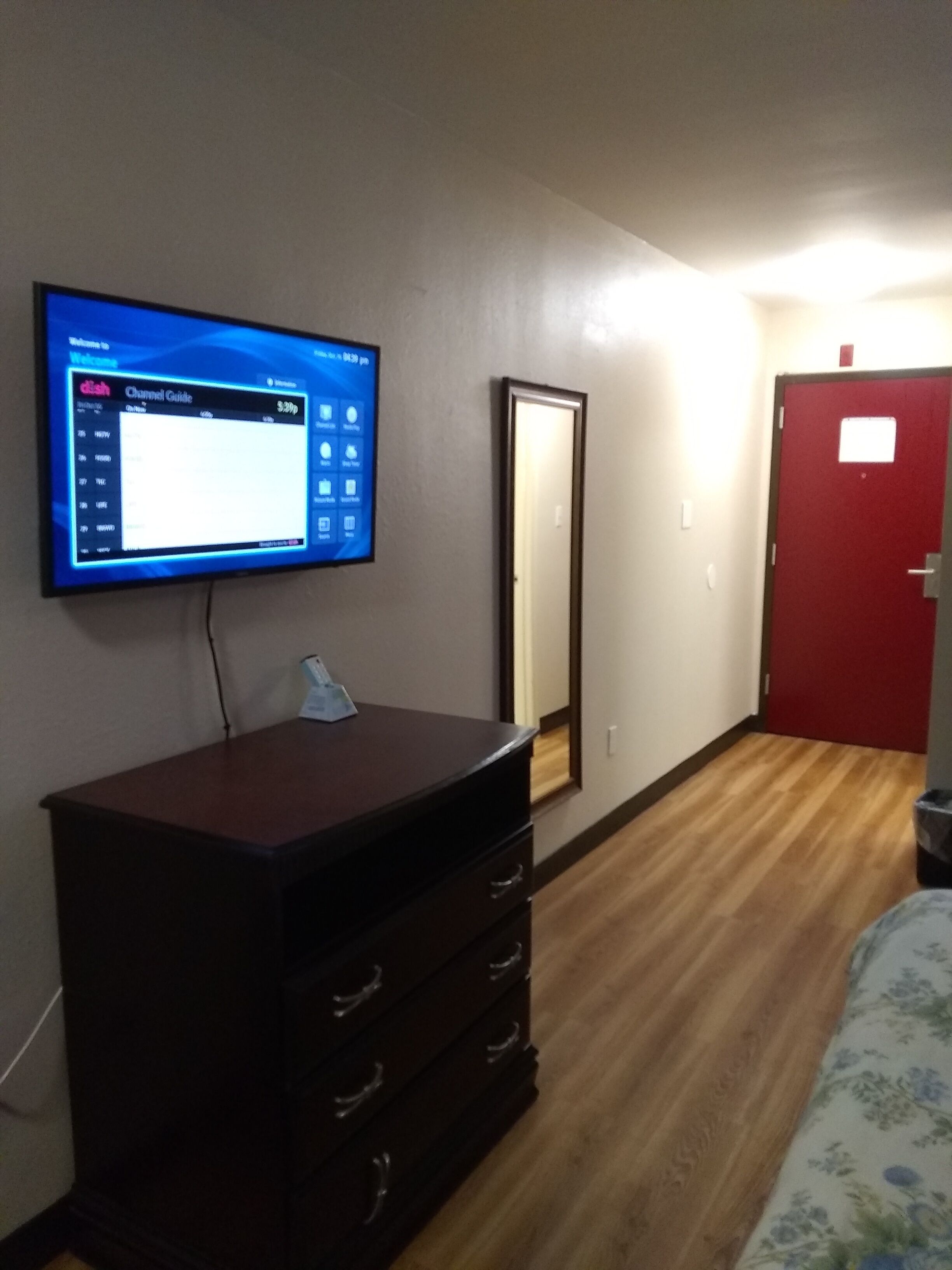 A hotel room with a flat-screen TV mounted on the wall displaying a channel guide, a wooden dresser with drawers, a mirror, and a red door.