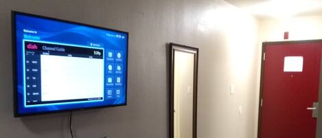 A hotel room with a flat-screen TV mounted on the wall displaying a channel guide, a wooden dresser with drawers, a mirror, and a red door.