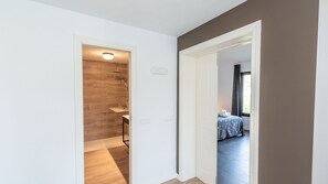 Un couloir menant à une salle de bain équipée d’une douche et d’un lavabo, ainsi qu’une chambre avec un lit et une fenêtre.