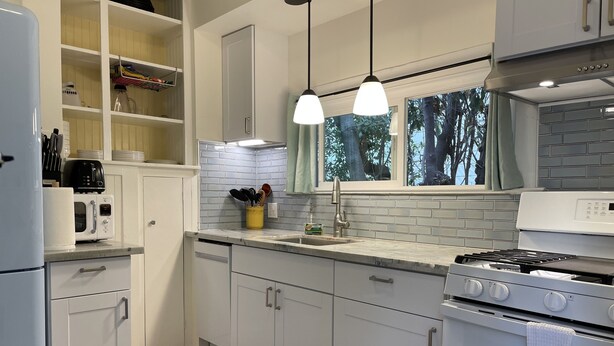 Une cuisine moderne avec des armoires blanches, un four encastré, un four à micro-ondes et une cuisinière à gaz. On y trouve des luminaires suspendus et une fenêtre donnant sur les arbres.