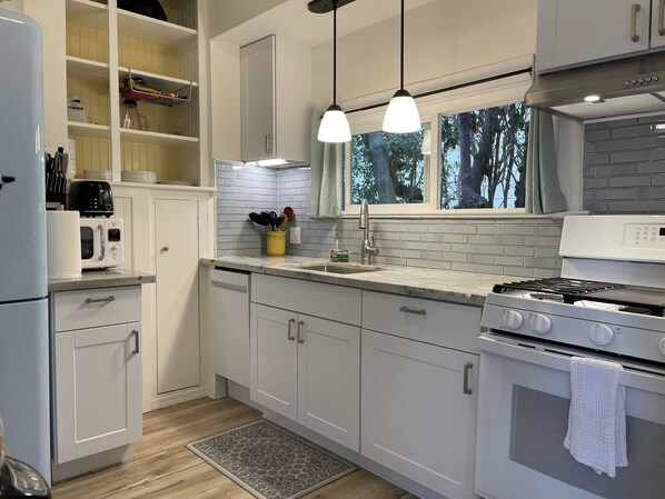 A modern kitchen with white cabinets, a built-in oven, a microwave, and a gas stove. There are hanging pendant lights and a window with a view of trees. - Darling Downtown, Walk Everywhere, Single Level, Quiet, AC, Pet Friendly (Santa Barbara)