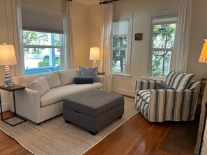 A living room with a sofa, ottoman, and armchair, two lamps, and a view of the outdoors.