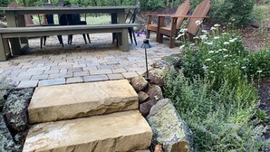 A stone pathway leading to a wooden bench and chairs in a garden setting.