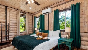 A wooden cabin bedroom with a bed, green curtains, a nightstand, and a window with a view of greenery. - New Luxury Cabin w/ King Beds, Game Room and Fire Pit  (Eureka Springs)