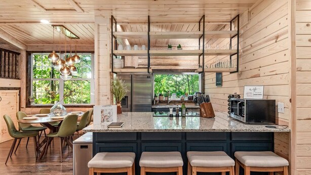 A modern kitchen with wooden cabinetry, a central island with stools, and a view of the outdoors. - New Luxury Cabin w/ King Beds, Game Room and Fire Pit  (Eureka Springs)