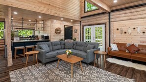 A spacious living area with a large gray sectional sofa, a wooden coffee table, and a patterned rug. There are two small wooden side tables and a brown leather sofa. The room features exposed wooden ceilings and walls, a large window with a view of greenery, and a kitchen area with a microwave and cabinets. - New Luxury Cabin w/ King Beds, Game Room and Fire Pit  (Eureka Springs)