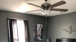A bedroom with a large bed, a ceiling fan, a nightstand, a lamp, and a painting on the wall.
