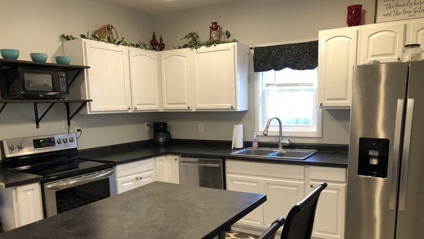 A kitchen with white cabinets, a stainless steel refrigerator, a black countertop, and a window with a black valance.