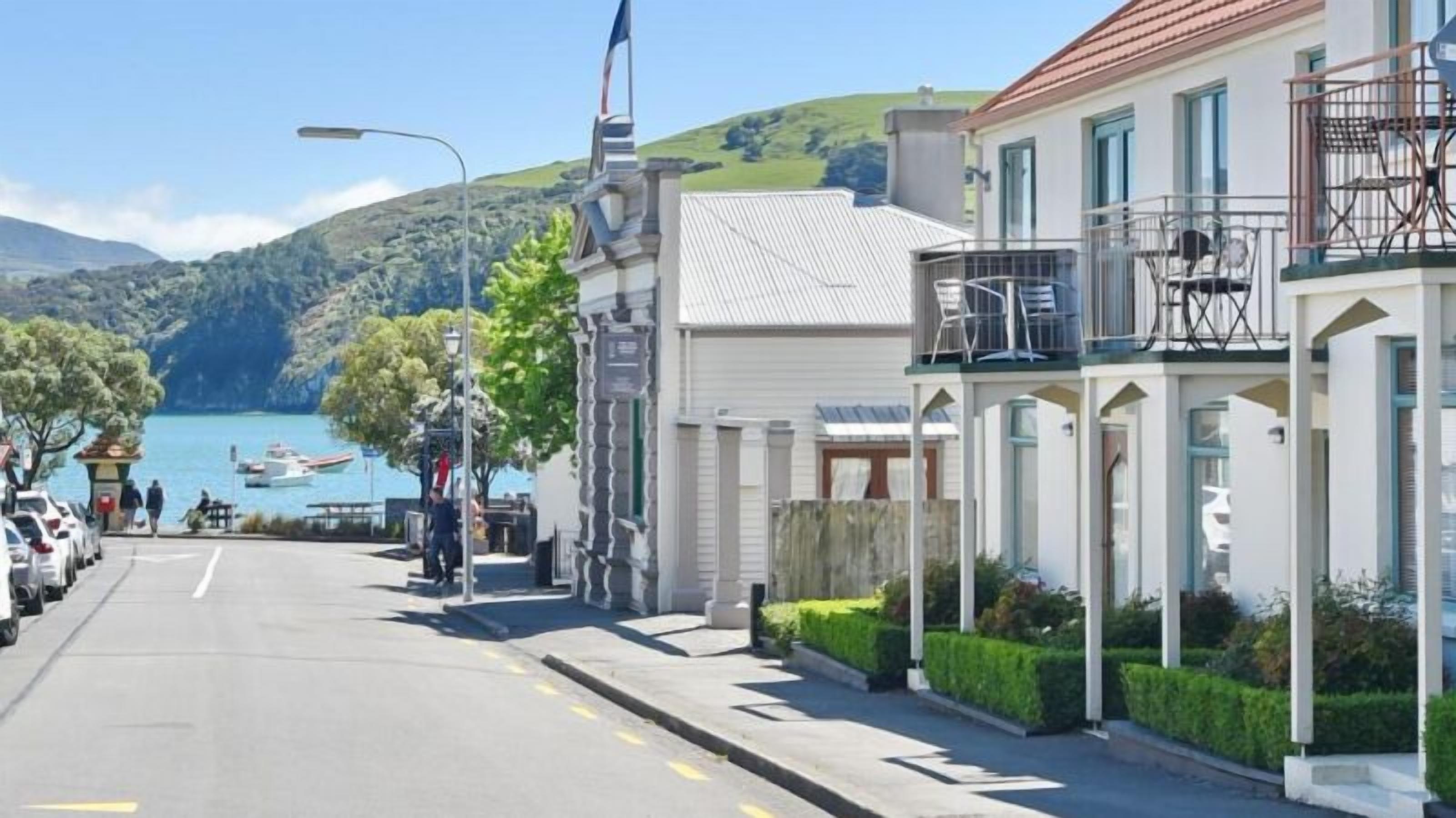 A coastal street with a row of buildings, a clear view of the sea, and parked cars along the road.