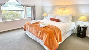 A hotel room with a large bed, two bedside lamps, a window with a view of buildings, and a small table with a lamp. - Central Akaroa Retreat - Balcony & Parking -Tumeke Akaroa (Akaroa)