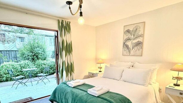 A hotel room with a bed, green bedspread, white pillows, two bedside lamps, a framed artwork on the wall, and a view of the outside through a sliding door. - Central Akaroa Retreat - Balcony & Parking -Tumeke Akaroa (Akaroa)