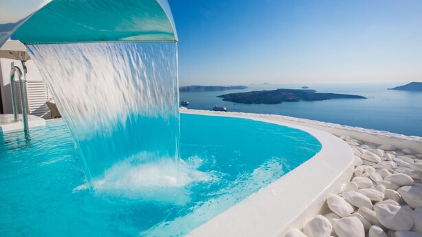 A pool with a water feature, surrounded by white stones and overlooking a coastal landscape. - Chic Hotel Santorini (Santorini)