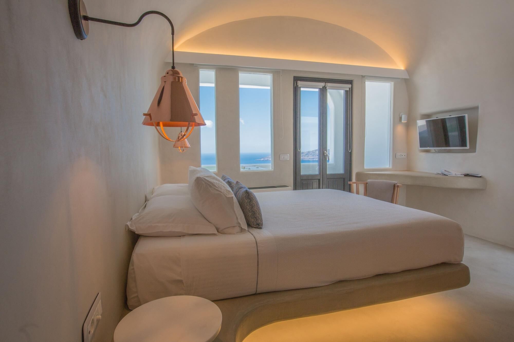 A modern bedroom with a large bed, a small round table, a TV, and a balcony with a view.