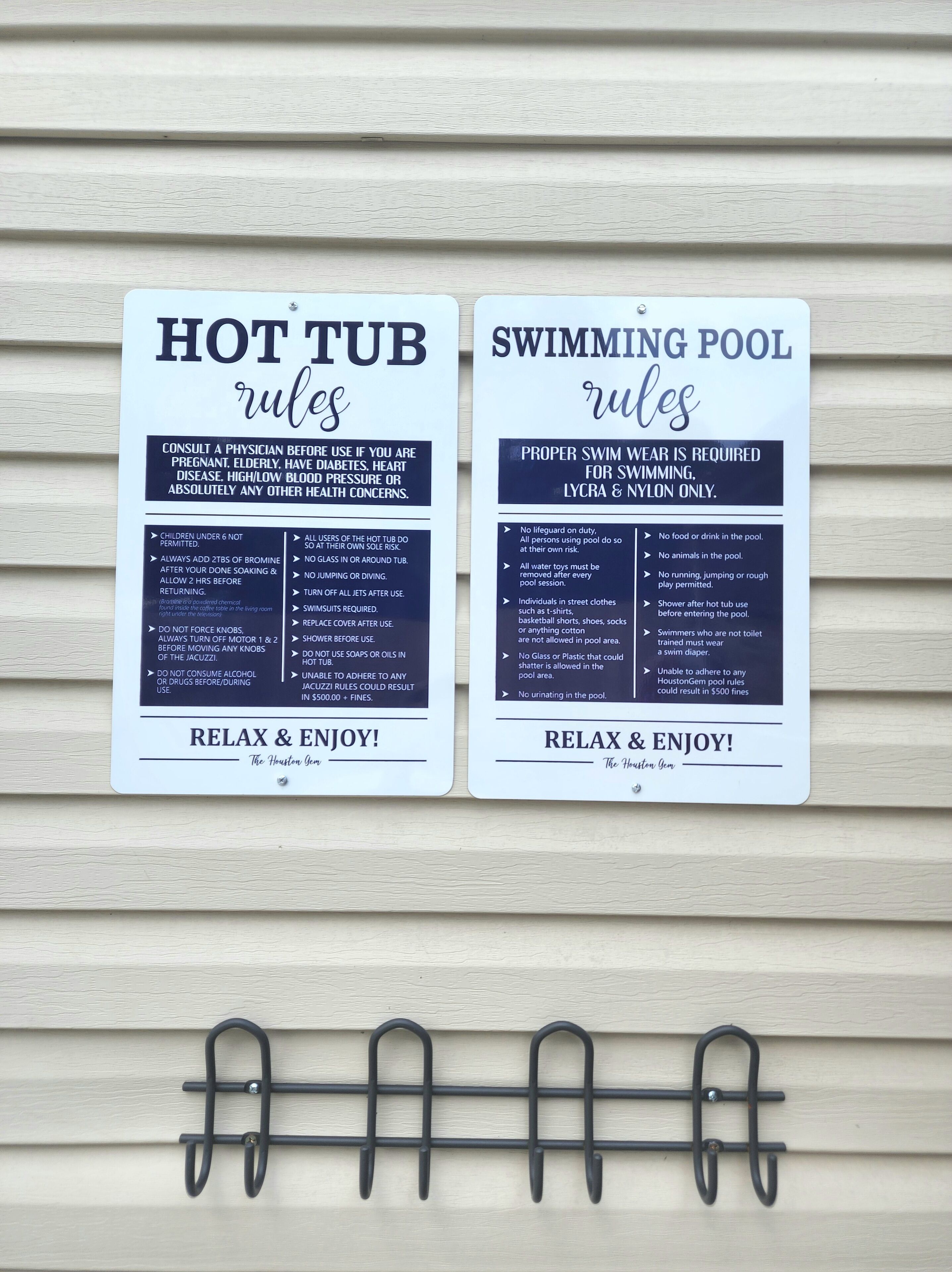 Two signs with rules for a hot tub and a swimming pool, mounted on a wall with a metal rack below.