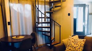 Ein kompakter Wohnbereich mit Wendeltreppe, einem Esstisch mit Stühlen, einem Sofa mit gemustertem Kissen und einem Couchtisch mit Fernbedienungen.