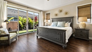 A bedroom with a large bed, a ceiling fan, a nightstand, a chair, and a view of a garden through a sliding glass door.