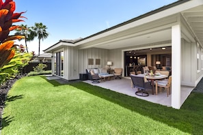 Una casa moderna con un patio cubierto, césped y una zona de comedor con mobiliario de exterior.