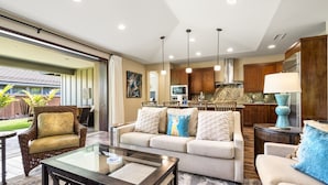 A spacious living room with a sofa, armchairs, and a glass coffee table. A kitchen with wooden cabinets and a refrigerator is visible in the background.