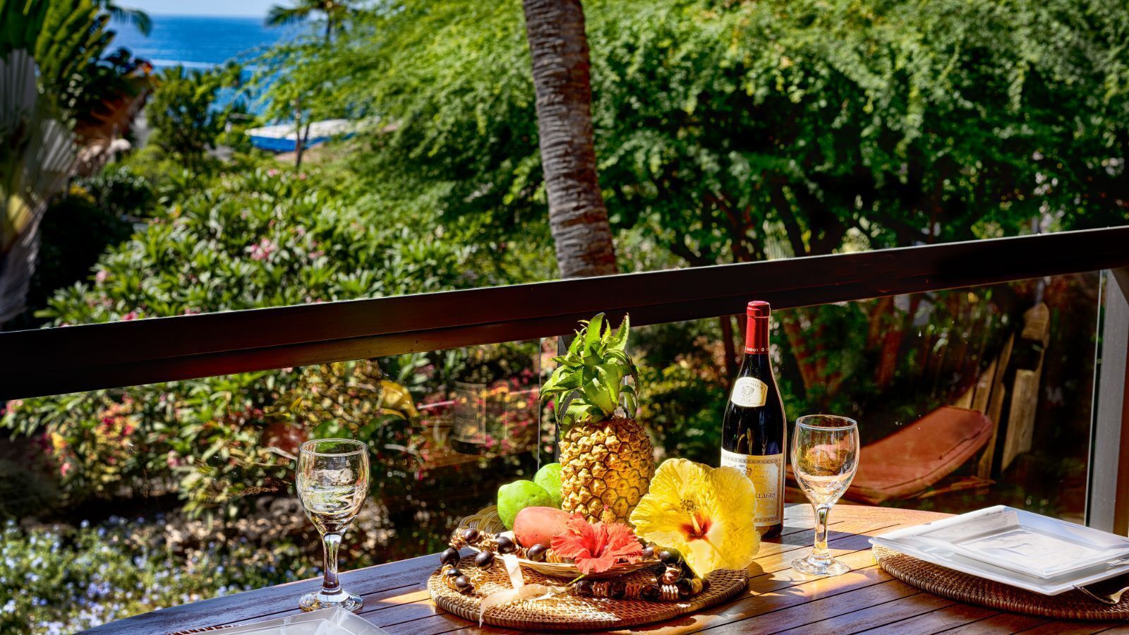 A table set with a pineapple, a bottle of wine, glasses, and tropical fruits, overlooking a lush garden and the ocean.