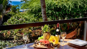 Una mesa servida con piña, botella de vino, copas y frutas tropicales, con vistas a un jardín exuberante y el océano.