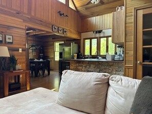 Un intérieur de chalet chaleureux avec une cheminée en pierre, des murs en bois, un coin repas et une cuisine équipée d’un réfrigérateur et offrant une vue sur le paysage par la fenêtre.