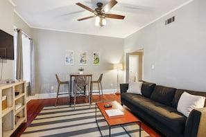 A living room with a ceiling fan, a sofa, a coffee table, a TV, and a dining area with chairs and a table. - Historic Savannah Garden Apartment on Jones Street (Savannah)