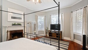 Un dormitorio con cama con dosel, chimenea, televisor sobre un atril de madera y un gran ventanal con cortinas.