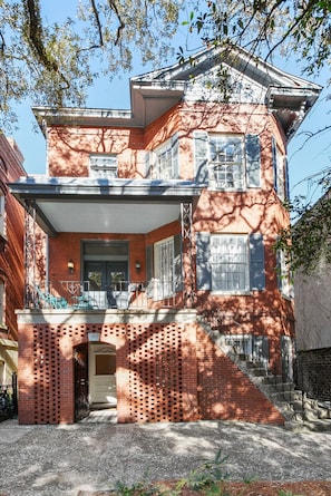 A two-story brick house with a covered front porch, a small front yard, and a staircase leading to the second floor. - Historic Savannah Garden Apartment on Jones Street (Savannah)