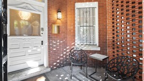 Fachada de ladrillo con una puerta blanca, una ventana con rejas decorativas y dos sillas de metal.