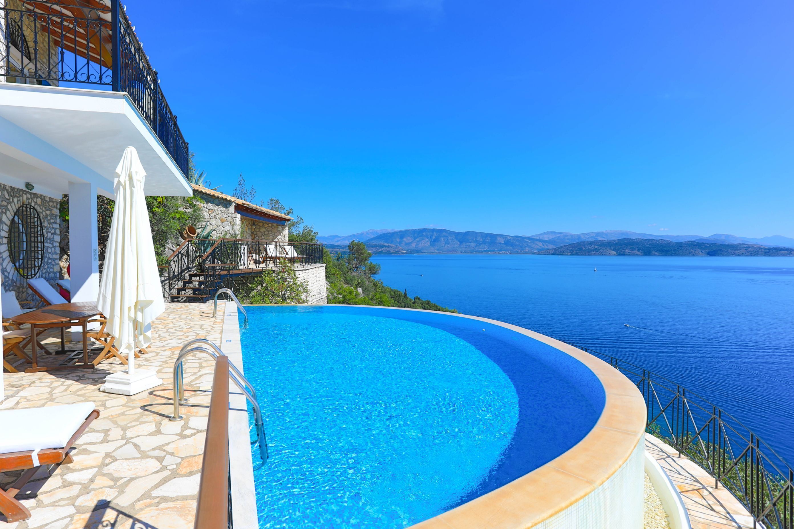 A pool with a curved edge, stone flooring, and a view of the sea and mountains.