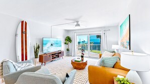 A modern living room with a surfboard, a TV, a sofa, and a view of the ocean. - Stylish Beachfront Condo – Balcony, Heated Pool & Steps to Pier (Cocoa Beach)
