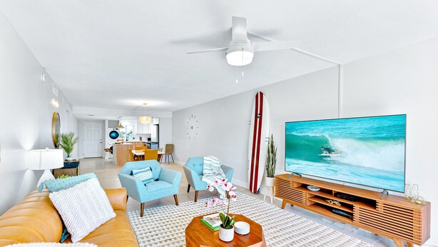 A modern living room with a large flat-screen TV, a ceiling fan, and a kitchen in the background. - Stylish Beachfront Condo – Balcony, Heated Pool & Steps to Pier (Cocoa Beach)