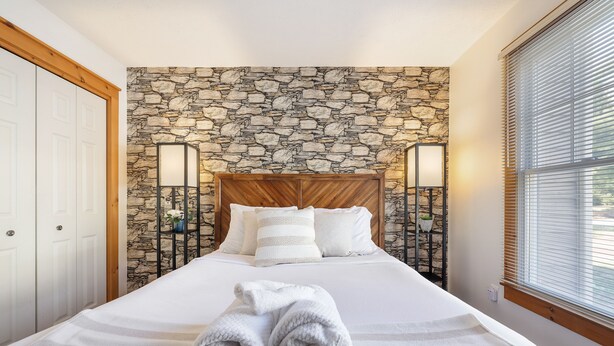 Una cama bien tendida con un fondo de pared de piedra, un cabecero de madera y dos lámparas de noche.