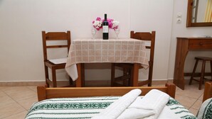 A neatly made bed with a patterned bedspread, a table with a checkered tablecloth, two chairs, a bottle of wine, and a vase with flowers. - Enjoy Villas (Santorini)