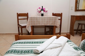 A neatly made bed with a patterned bedspread, a table with a checkered tablecloth, two chairs, a bottle of wine, and a vase with flowers. - Enjoy Villas (Santorini)