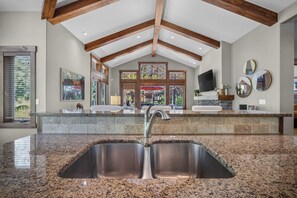 A kitchen with granite countertops, a stainless steel sink, wooden beams, and a stone backsplash.