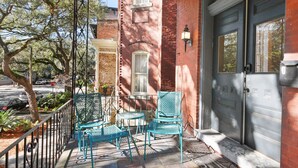A covered porch with two chairs and a small table, a brick building, and a dark-colored door. - Renovated Jones Street Parlor Apt +Private Parking (Savannah)