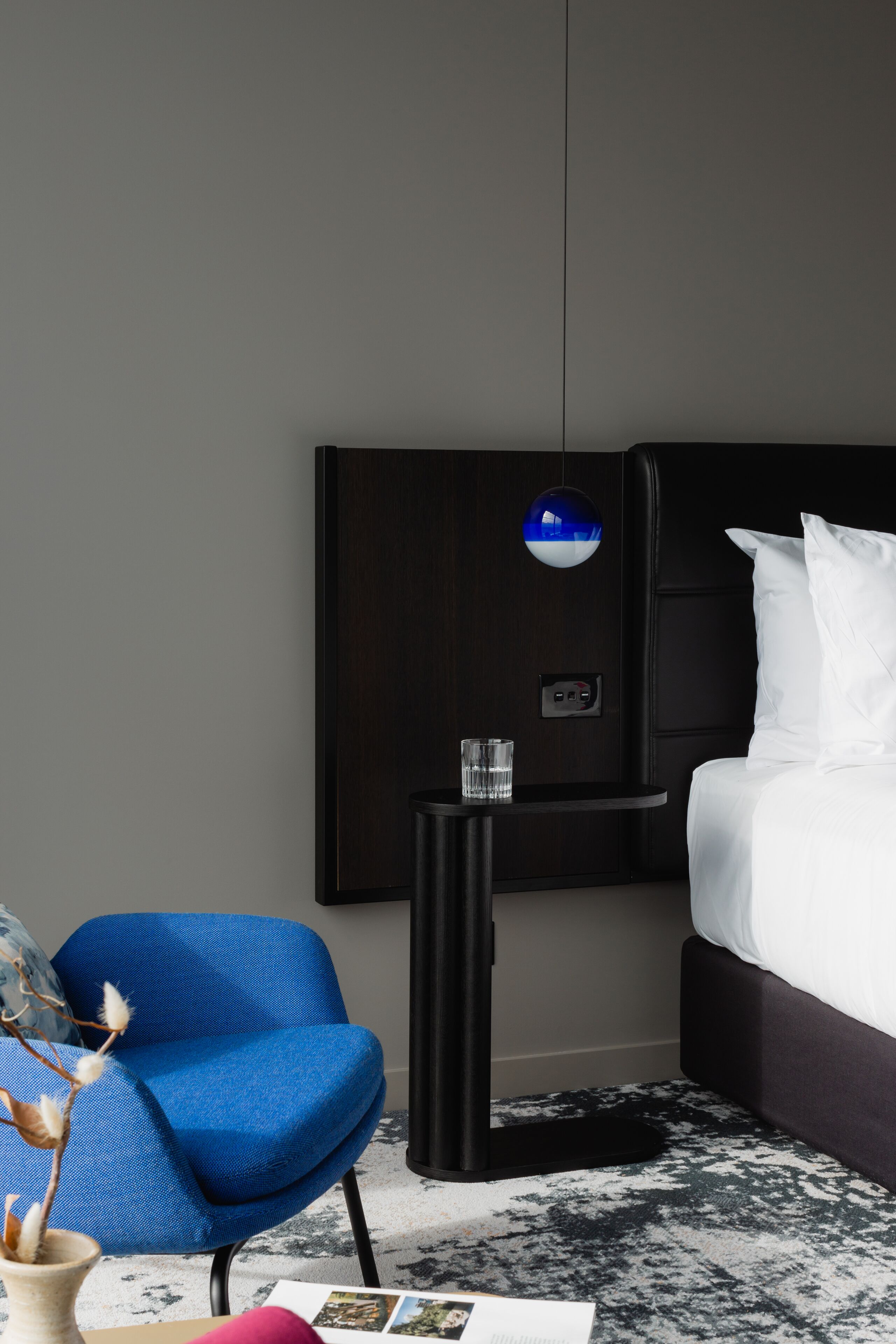 Een moderne hotelkamer met een hoofdeinde van donker hout, een blauwe fauteuil, een nachtkastje met een glas en een wit bed.