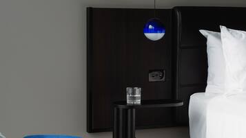 Een moderne hotelkamer met een hoofdeinde van donker hout, een blauwe fauteuil, een nachtkastje met een glas en een wit bed.