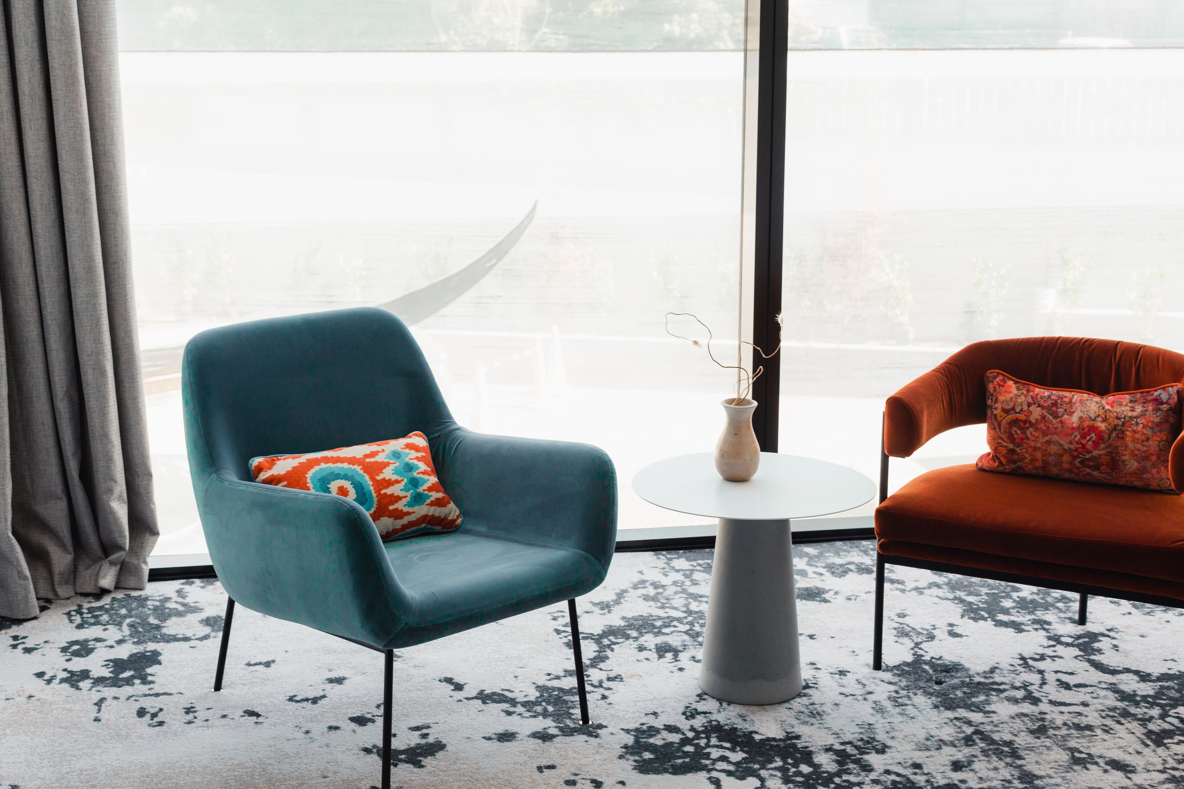 Une pièce avec deux chaises, une turquoise et une orange, chacune agrémentée d’un coussin à motifs. Une petite table blanche avec un vase est placée au centre. La pièce est dotée d’une grande fenêtre donnant sur un plan d’eau.