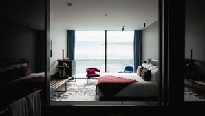 A modern hotel room with a large bed, a seating area with a red chair, and a view of the ocean through large windows. - Ebb-Dunedin (Dunedin)