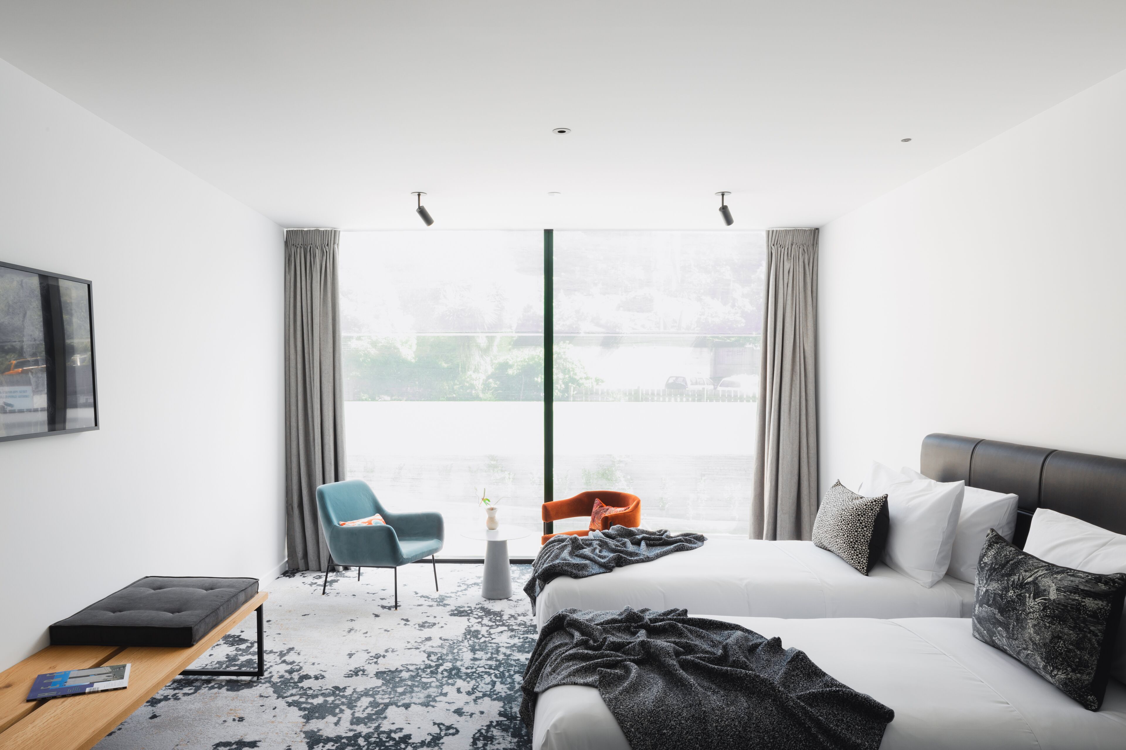 A modern hotel room with a large bed, a desk, a chair, and a view of the outdoors through a large window.