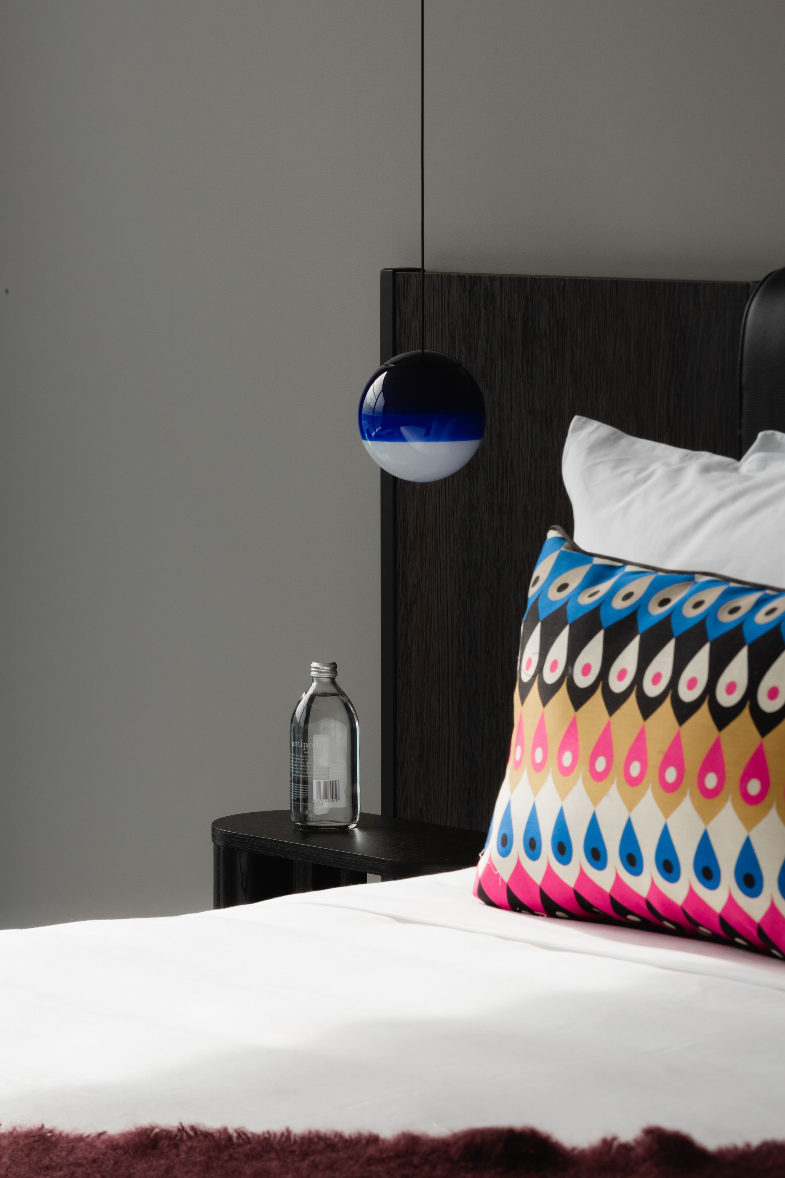 A neatly made bed with a patterned pillow, a bedside table with a bottle, and a wall-mounted lamp.