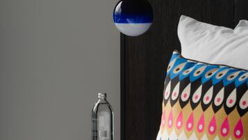 A neatly made bed with a patterned pillow, a bedside table with a bottle, and a wall-mounted lamp.