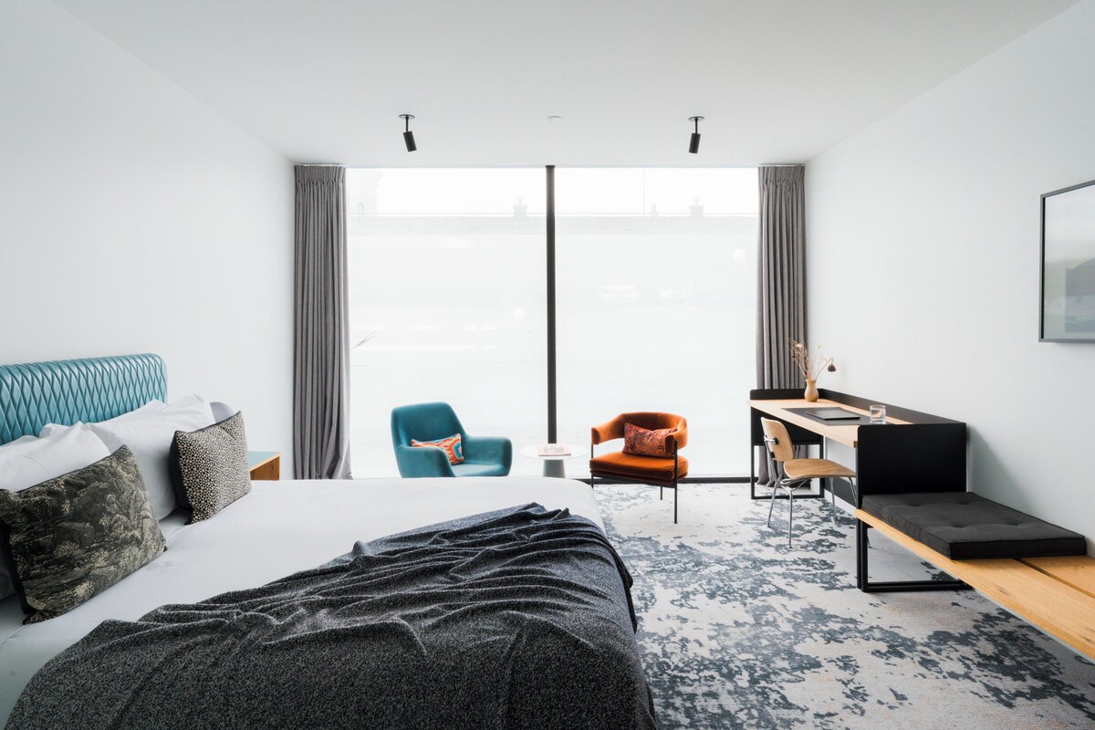 Une chambre d’hôtel moderne, dotée d’un grand lit, d’un bureau, de deux chaises et offrant une vue sur l’extérieur grâce à une grande fenêtre.