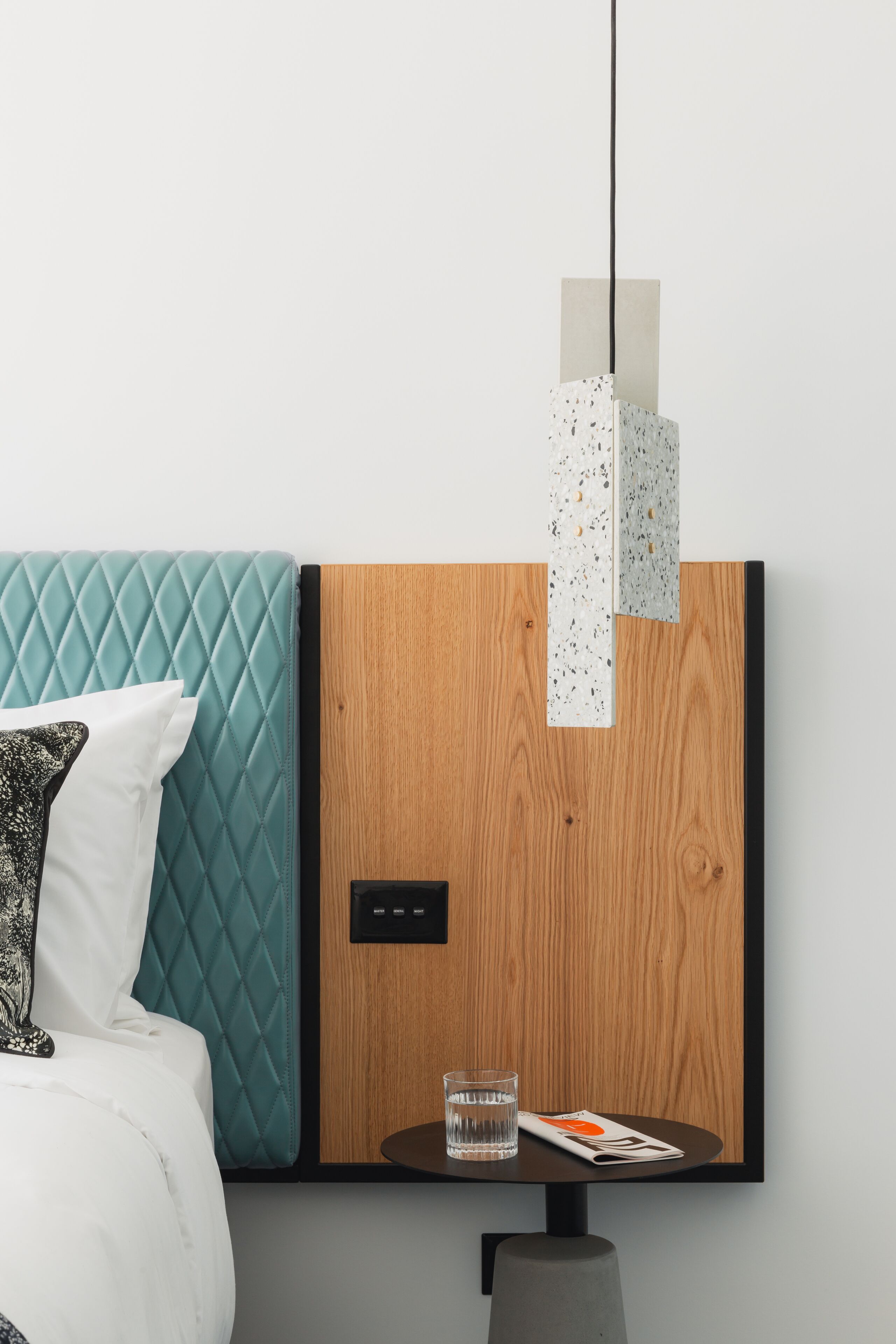 Un dormitorio moderno con un cabecero acolchado color turquesa, un panel de madera en la pared, una mesita de noche con un vaso de agua y una revista.
