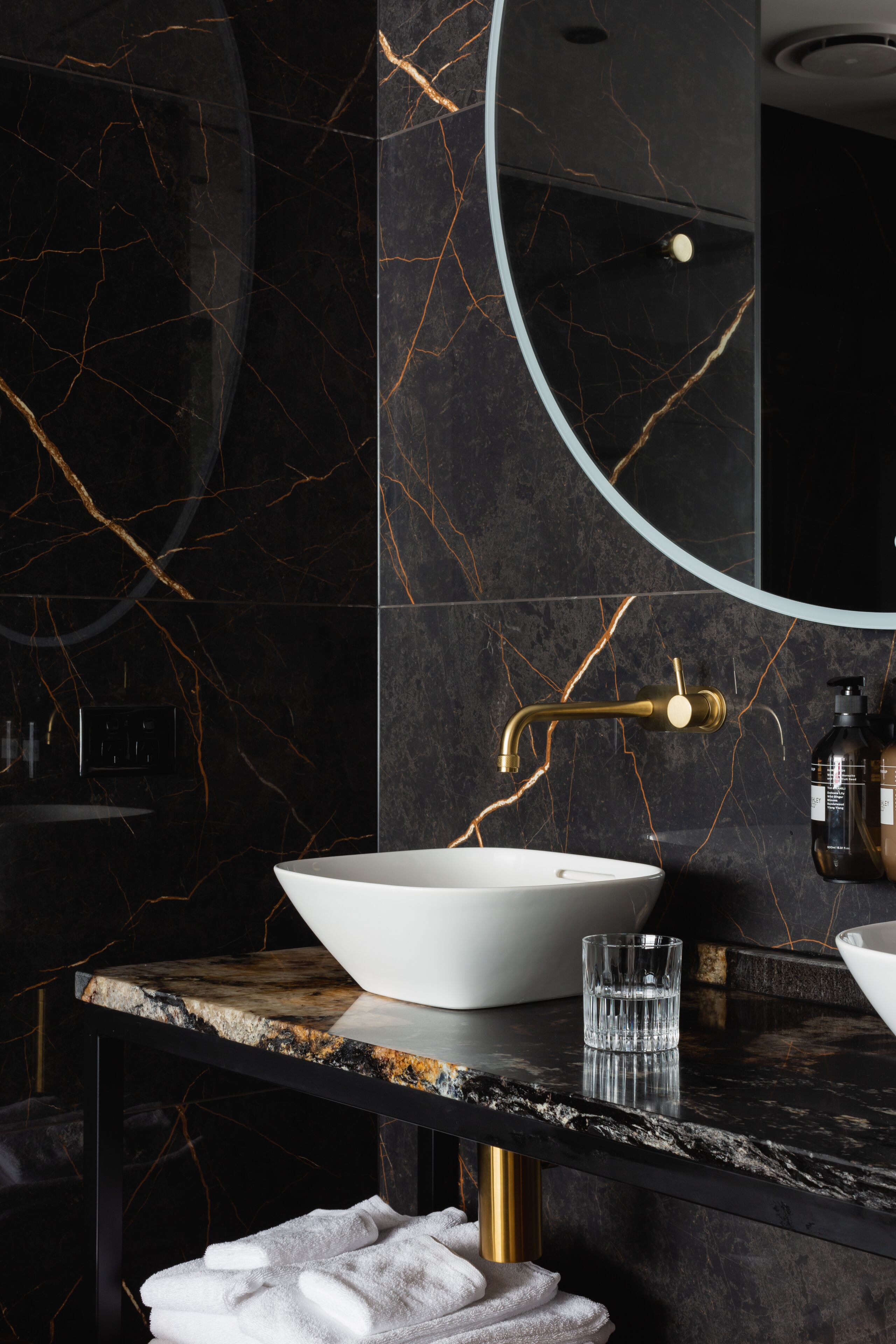 A modern bathroom with a white vessel sink, gold faucet, and a round mirror.